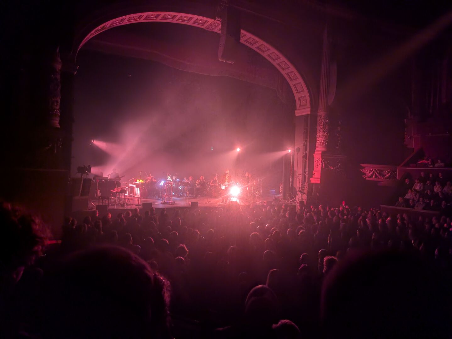 De zaal en het orkest tijdens een van de gespeelde nummers. Donkere zaal met rode verlichting en een tapijt van mensen.