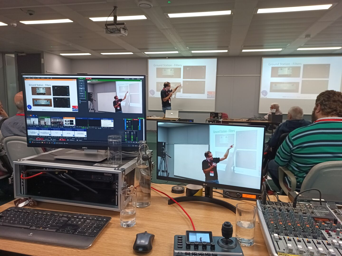 A speaker presenting in the background with video shown on various screens on a table with some gear