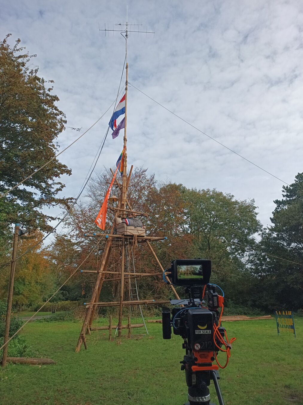 wooden tower built with big wooden poles and rope on a grass field. in the top, antennas are mounted. the mast is ~18m high. in tge foreground, a camera can be seen filming the mast