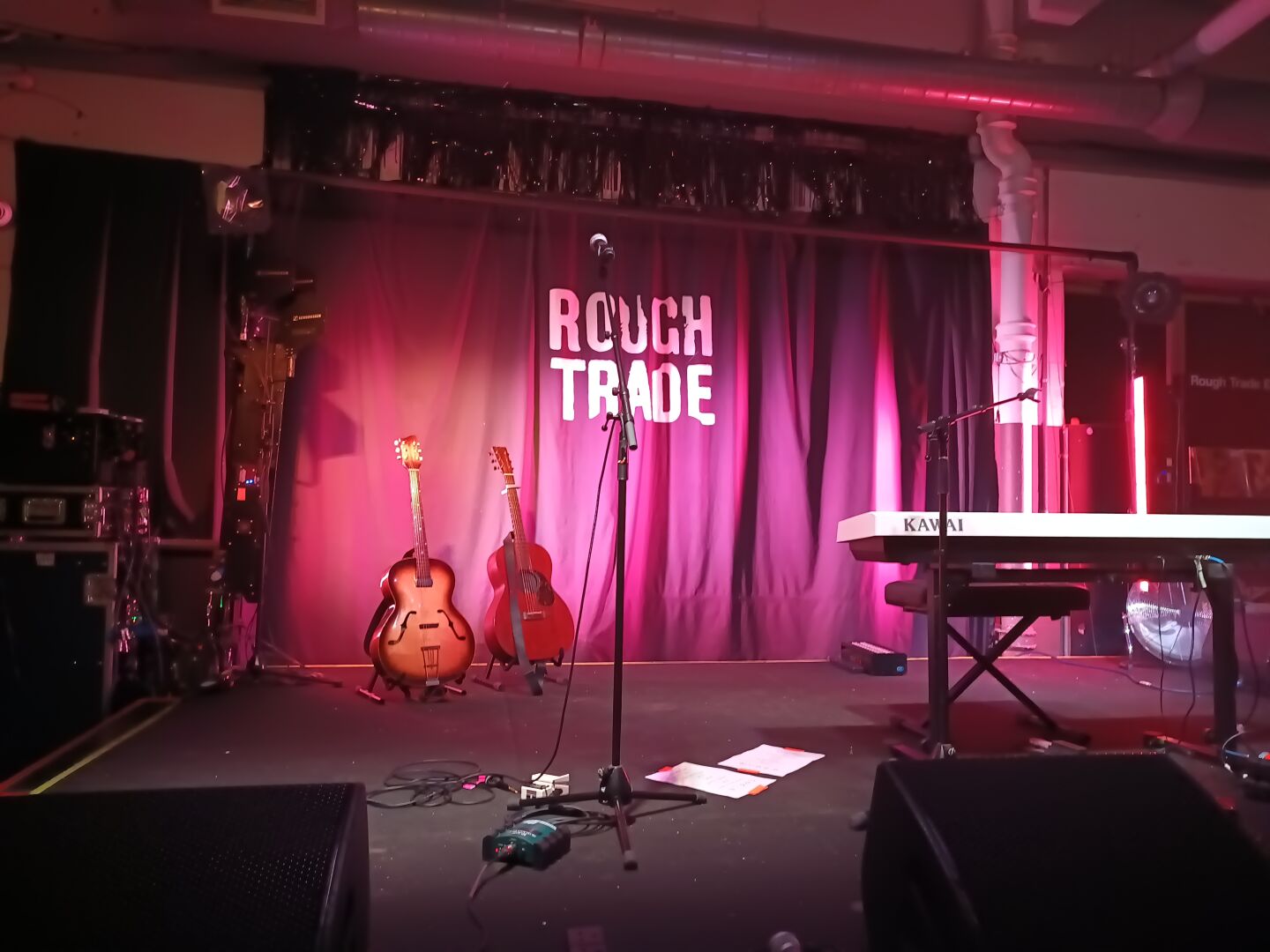 stage with rough trade flag in the background and pink coloured stage lights. a piano, two guitars and two microphones are present