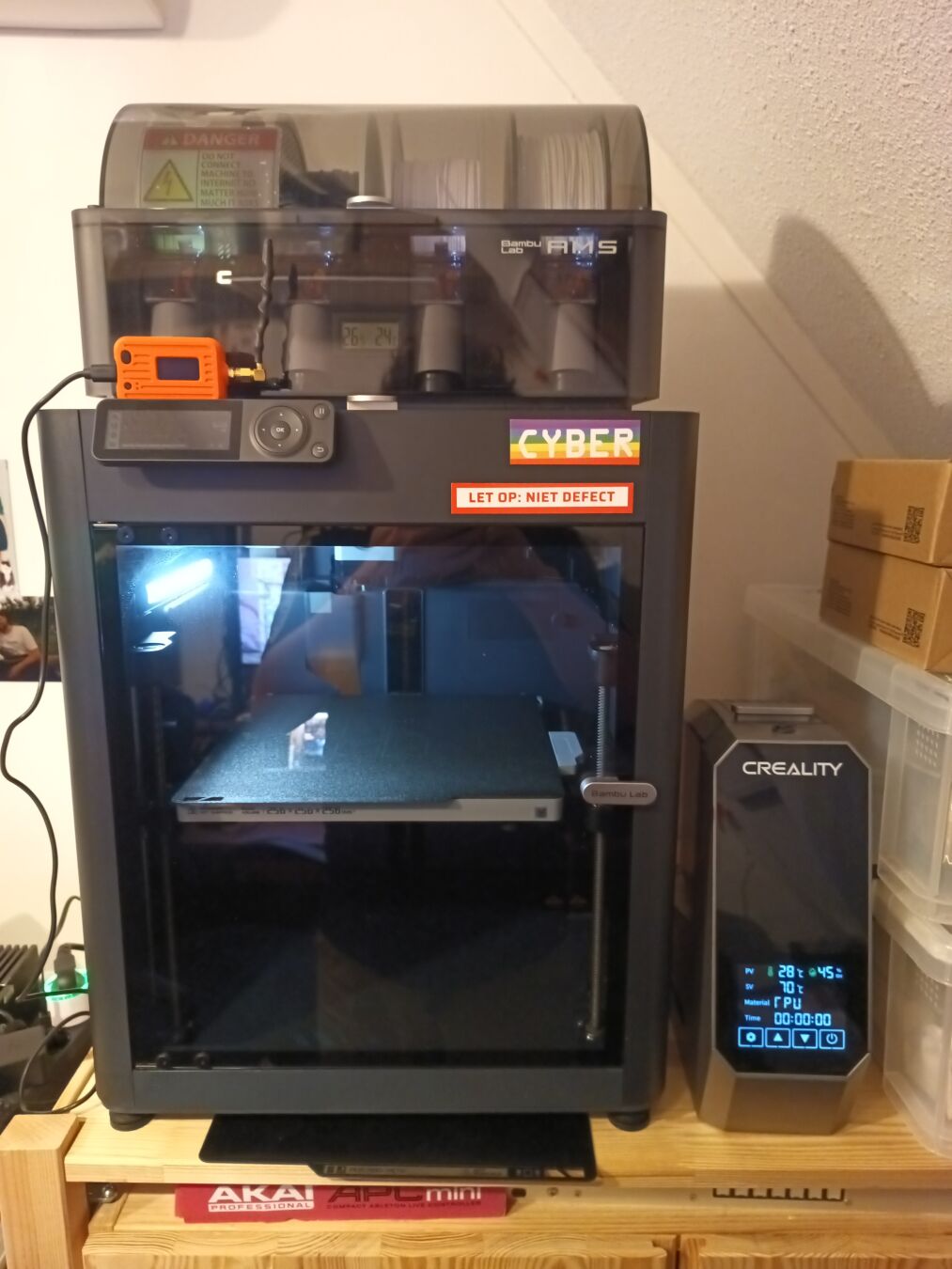 Bambu Labs P1S 3d printer with AMS material system. a filament dryer is visible on the right. on top of the printer, a 3d printed case can be seen containing a Heltec MeshCore node. the bambu labs logo is covered with a rainbow cyber sticker and a warning label stating "let op: niet defect", translation: attention: not broken