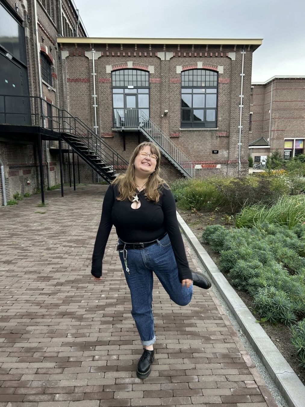Emily standing on one leg, the other leg bent backwards. Wearing blue jeans and a black top. The background is an industrial building from the 1900s