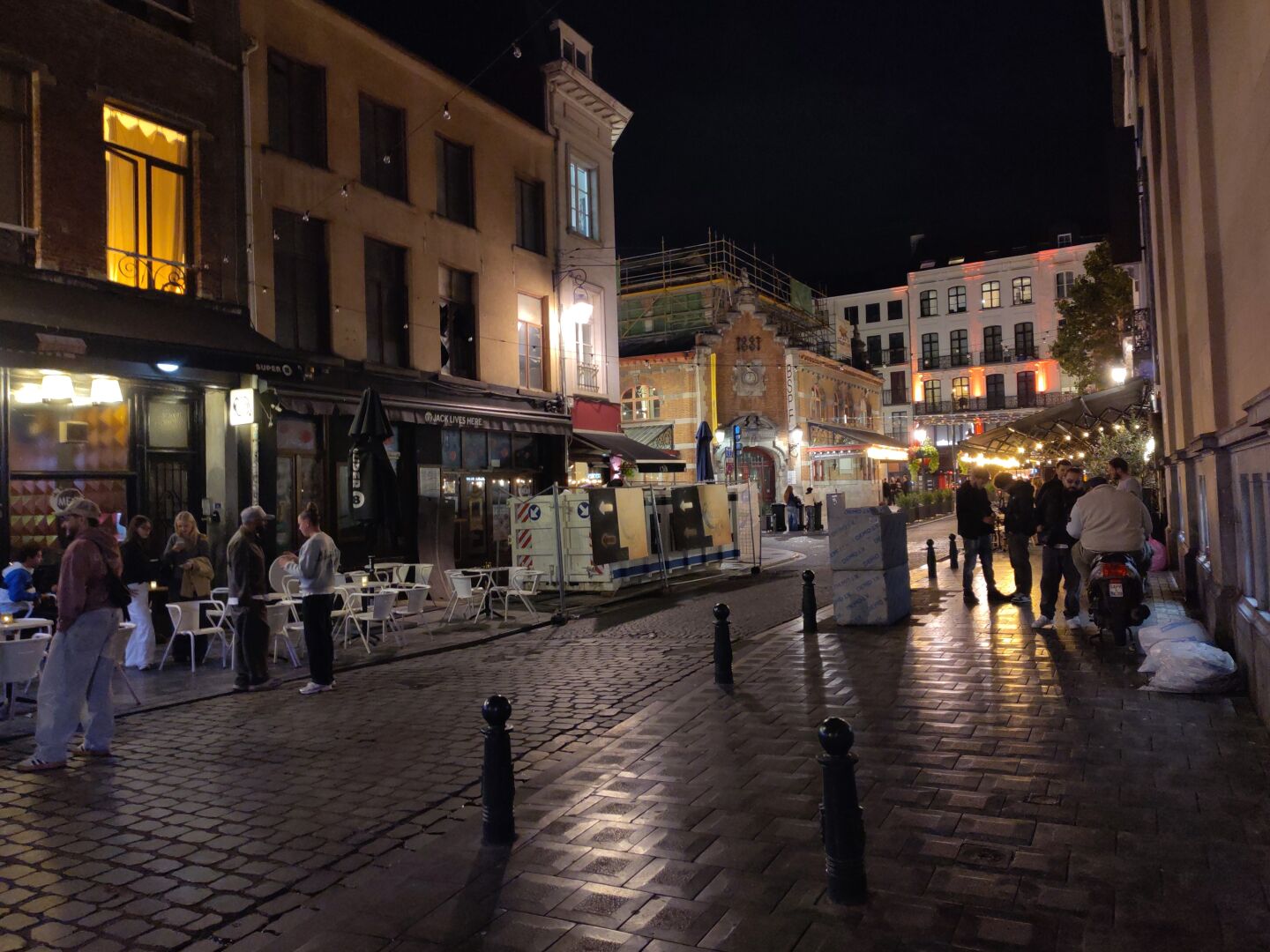 An evening street scene. You can see cafés with outdoor seating areas and people sitting or walking around.