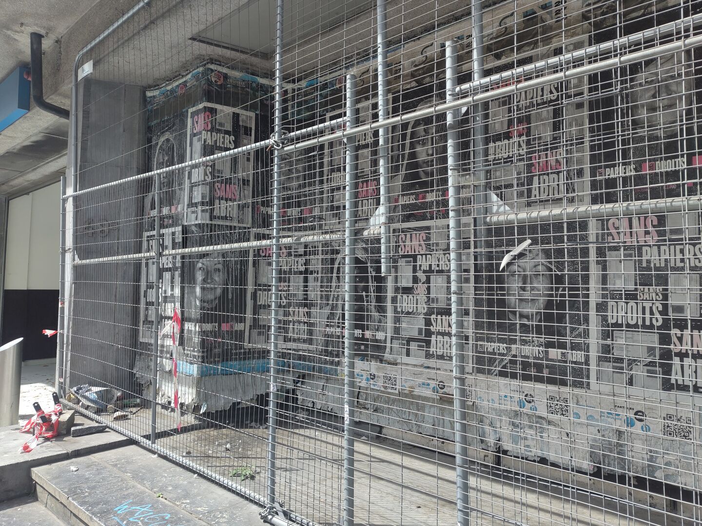A street-corner niche is blocked off by a chain-link fence, presumably to prevent people from taking shelter there. Behind the fence, on the wall, hang posters featuring portraits and the text 'Sans papiers, sans droits, sans abri'.
