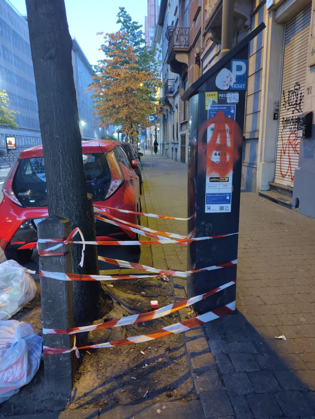 A red-and-white warning tape is wrapped haphazardly around a parking meter, a wooden post, and a tree trunk. There is no obvious purpose to it.
