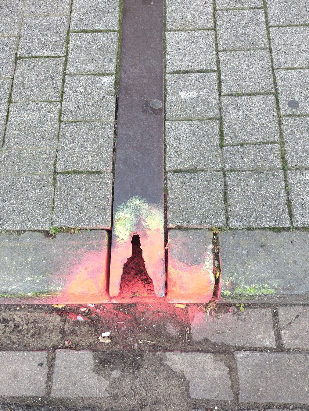 A metal gutter is embedded in a sidewalk. It is damaged at one end, where it has been spray-painted with orange safety paint.
